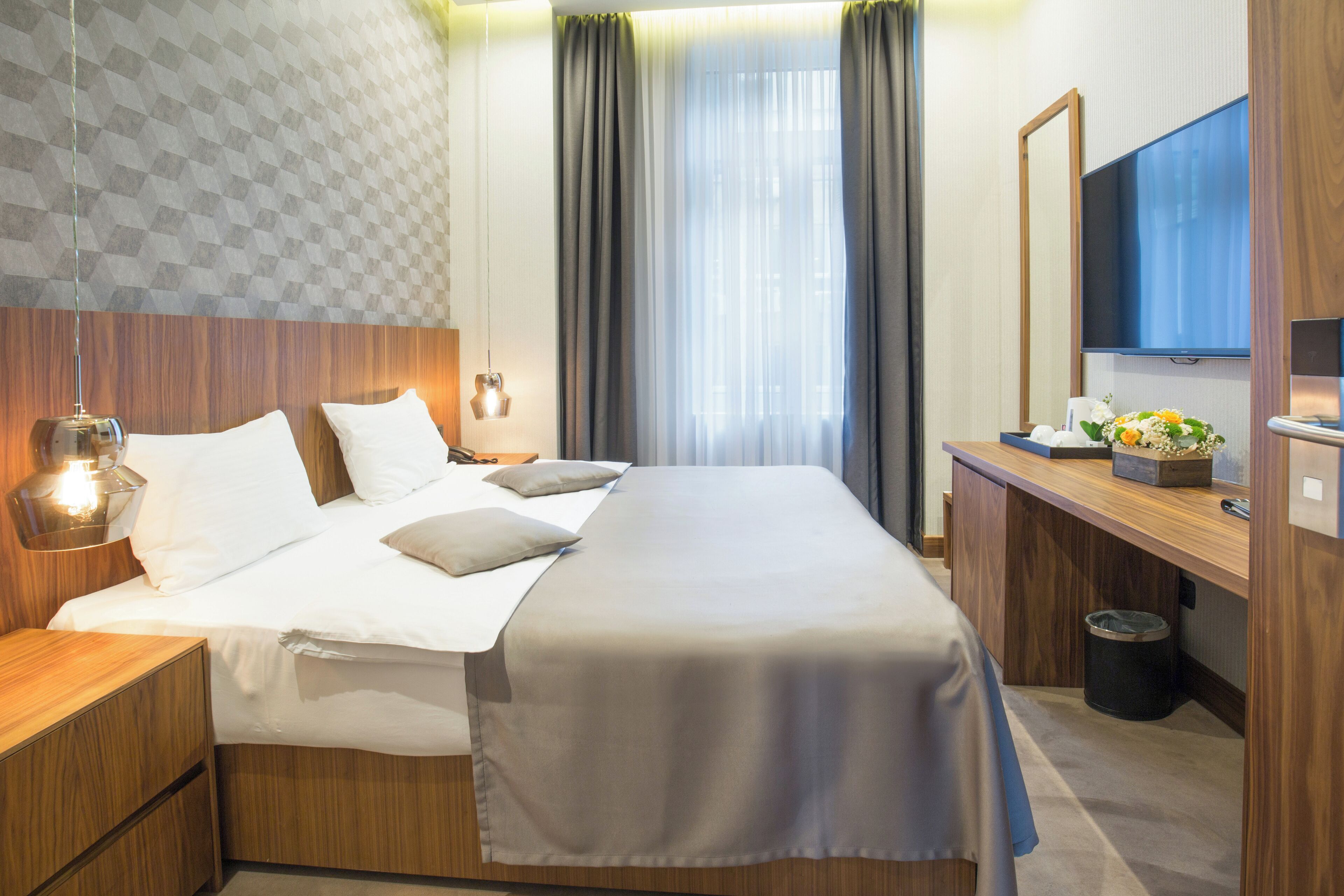 Ein Hotelzimmer mit Bett, Nachttischen, einem Schreibtisch mit Computer, einem Fernseher und einem Fenster mit Vorhängen.