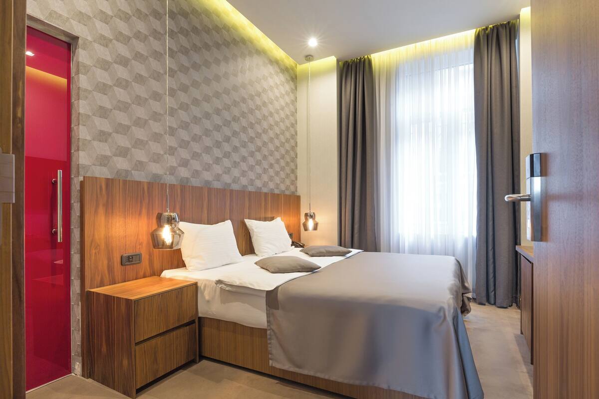 A modern hotel room with a large bed, wooden headboard, bedside tables, and a red door.