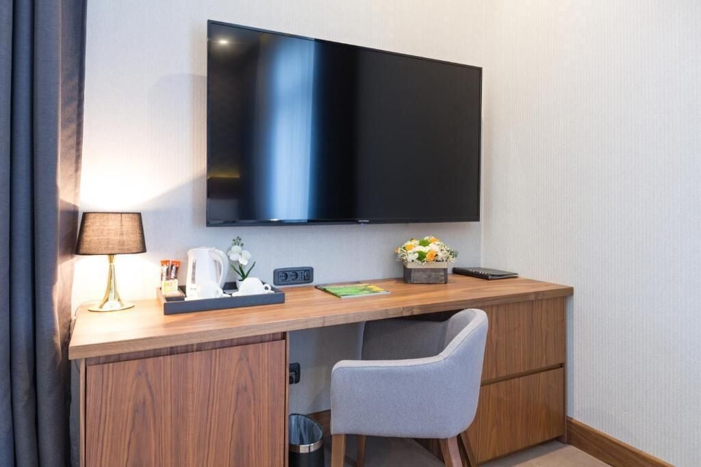 A hotel room with a wooden desk, a flat-screen TV, a lamp, a trash bin, and a grey chair.