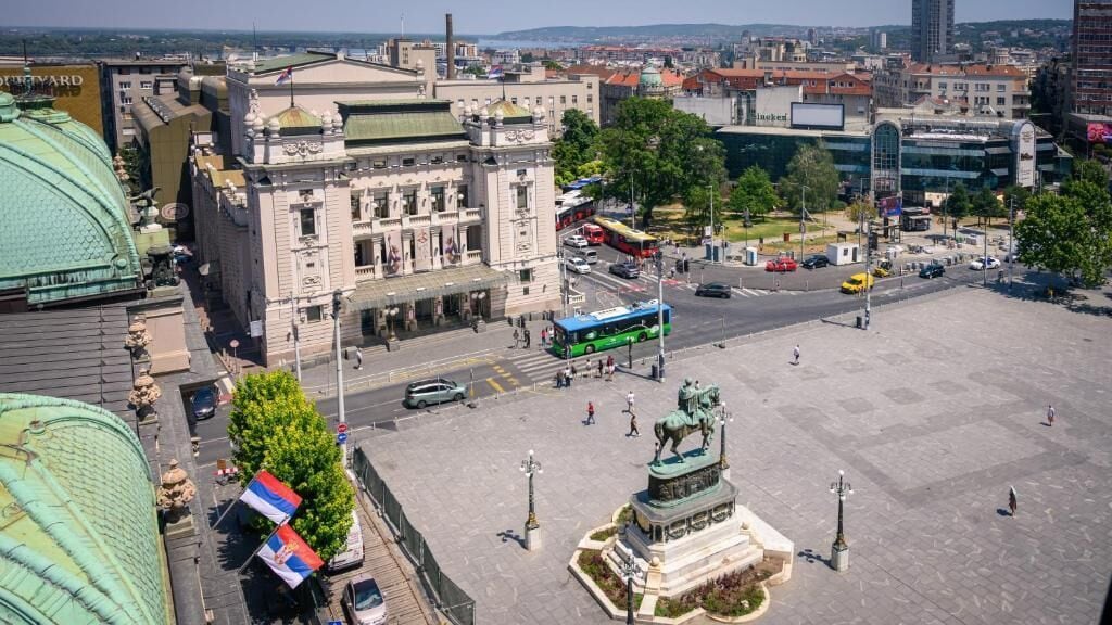 Ein Stadtplatz mit einem markanten Gebäude, einer Statue und einem grünen Bus im Vordergrund.