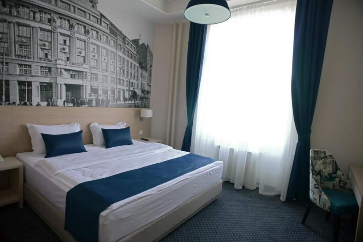 A hotel room with a large bed, a headboard with a cityscape mural, a desk with a chair, and a window with curtains.