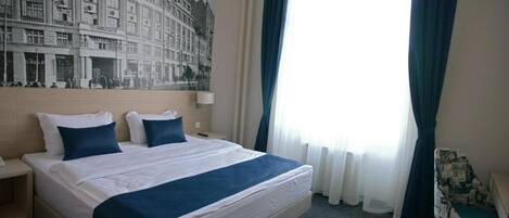 A hotel room with a large bed, a headboard with a cityscape mural, a desk with a chair, and a window with curtains.