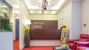 A reception area with a wooden reception desk, a chandelier, a red carpeted floor, a red leather sofa, and a potted plant. - Five Points Square - City Center (Belgrade)