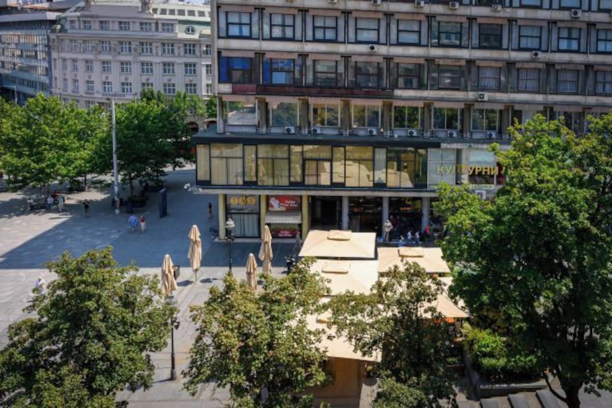 Egy többemeletes épület, melynek földszintjén üzlethelyiségek találhatók, és amelyet fák és egy közteret foglalnak körül.