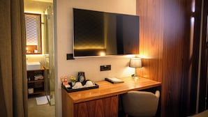 A hotel room with a wooden desk, a flat-screen TV mounted above it, a bedside lamp, a chair, a small shelf with toiletries, and a shower area visible through a glass door. - Five Points Square - City Center (Belgrade)