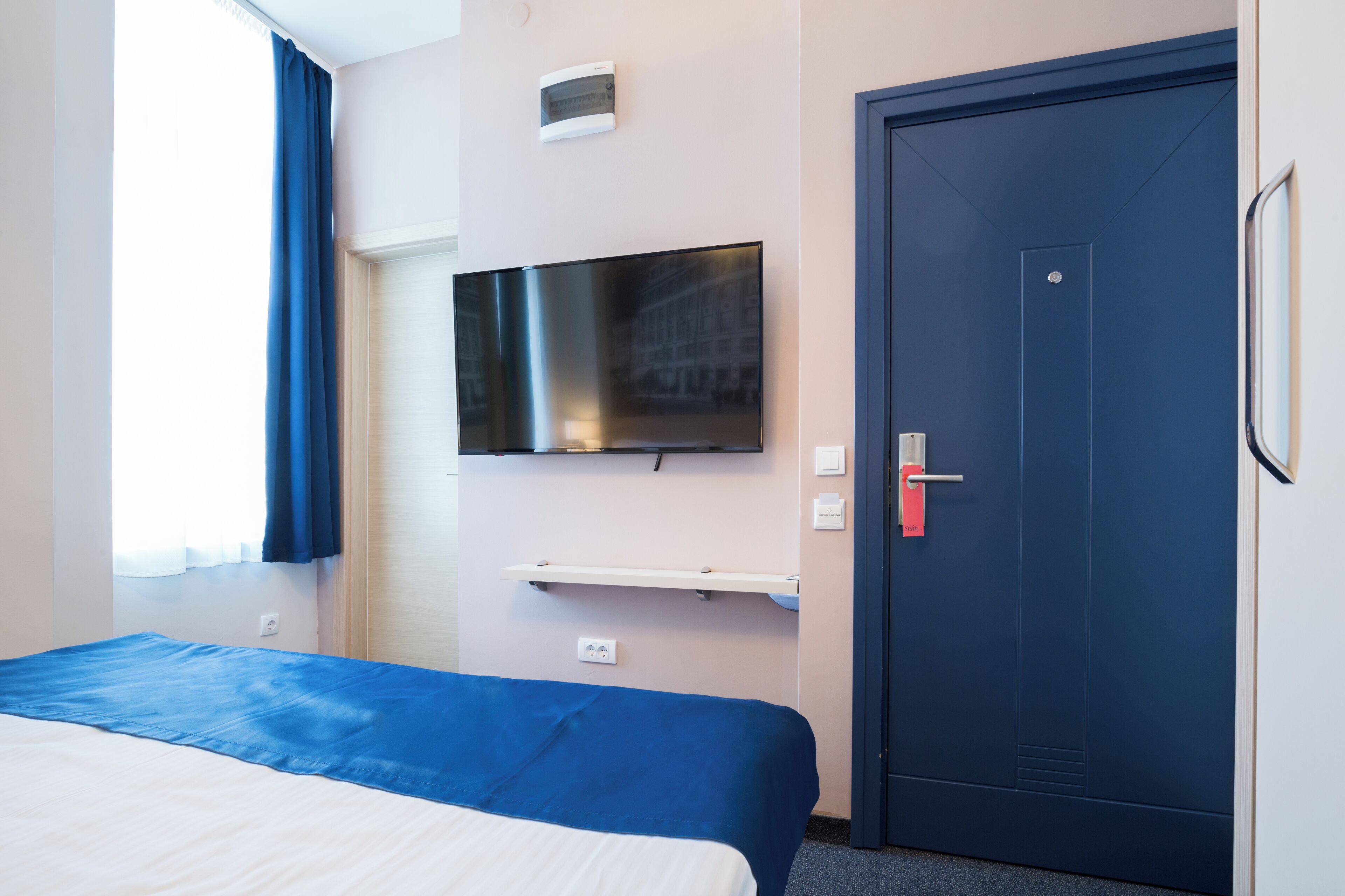 A hotel room with a bed, a flat-screen TV mounted on the wall, a door, and a window with curtains.