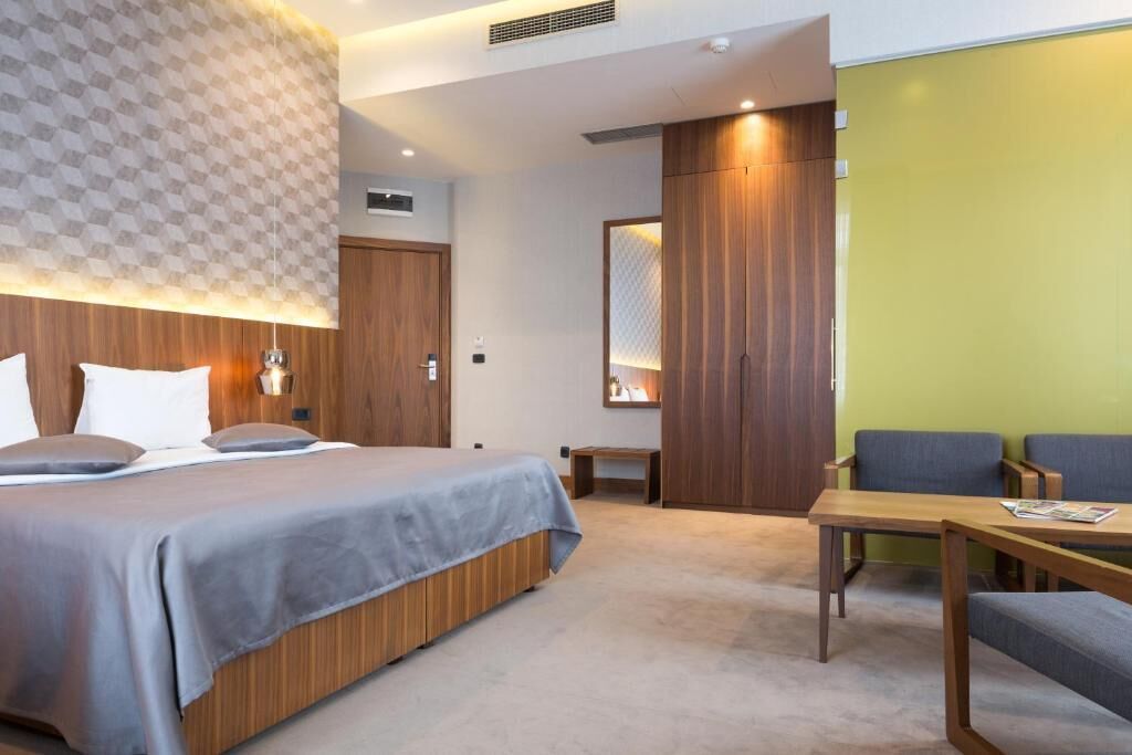 Une chambre d’hôtel moderne avec un grand lit, une tête de lit en bois, une petite table et un coin salon avec deux fauteuils.