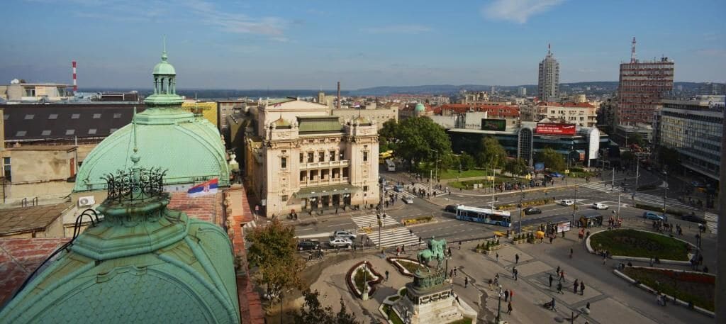 Eine Stadtansicht mit einer markanten grünen Kuppel, einem zentralen Platz mit einem Brunnen und umgebenden Gebäuden.
