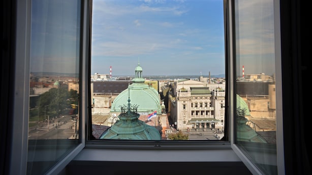 A view through a window of a cityscape with historical buildings and a prominent green dome structure. - Five Points Square - City Center (Belgrade)