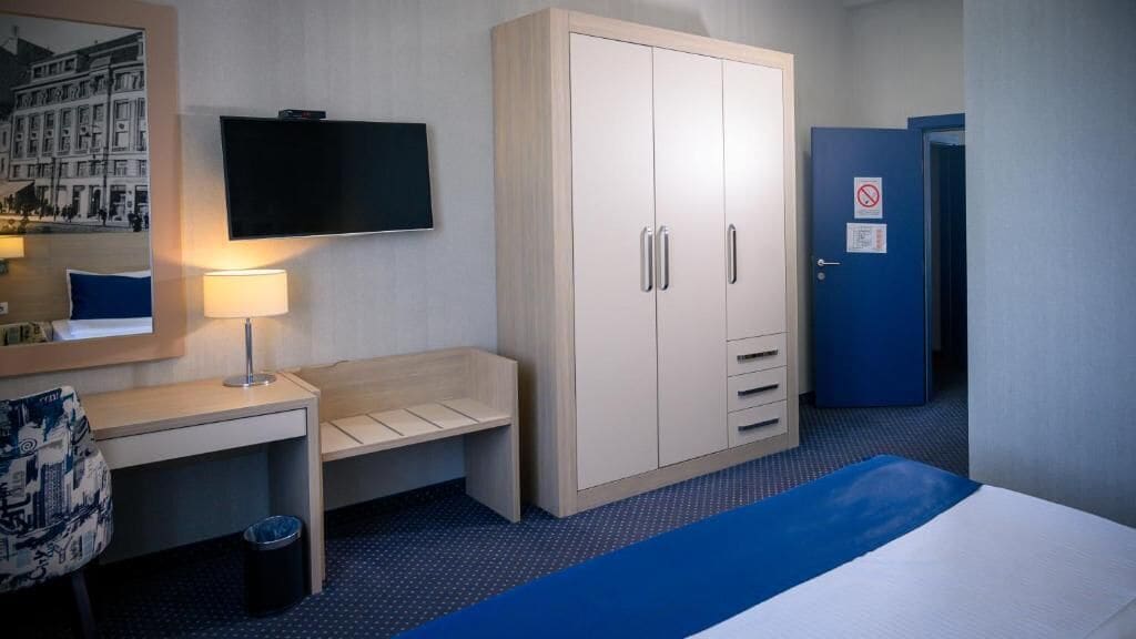 A hotel room with a bed, a desk, a chair, a wardrobe, a television, and a door with a 'no entry' sign.