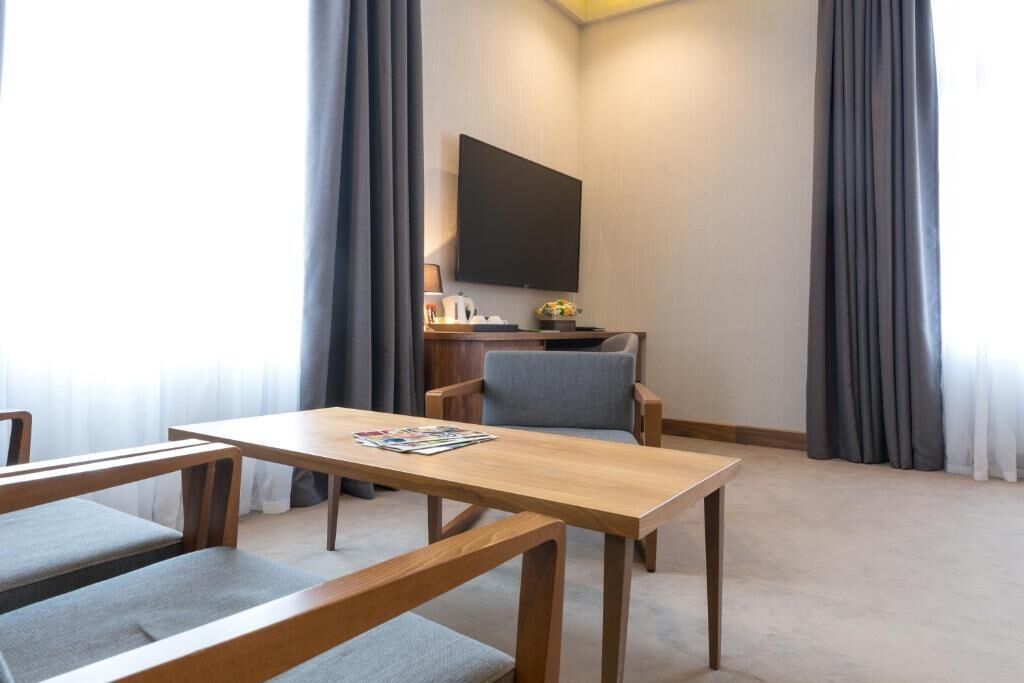 A hotel room with a wooden table, two grey cushioned chairs, a flat-screen TV mounted on the wall, and a small desk with a lamp and decorative items.