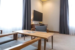 Quarto de hotel com mesa de madeira, duas cadeiras estofadas cinzentas, televisão de tela plana na parede e pequena escrivaninha com abajur e objetos decorativos.