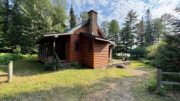 Une cabane rustique avec une cheminée, entourée d’une zone herbeuse et d’arbres.