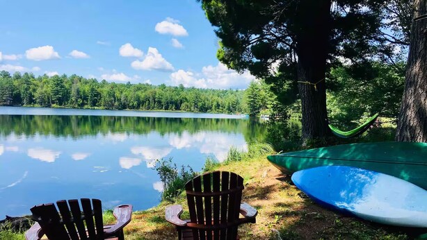 Two Adirondack chairs facing a calm lake with a hammock and a canoe nearby. - The Loon Call: Gorgeous Lakefront Family Cottage (Ladysmith)