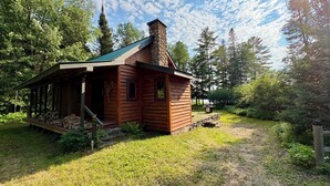 A rustic cabin with a chimney, a porch with stacked firewood, and a stone wall. - The Loon Call: Gorgeous Lakefront Family Cottage (Ladysmith)