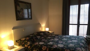 Et soveværelse med blomstret sengetæppe, to natborde med lamper, et spejl på væggen og et vindue med persienner.