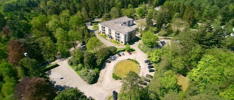 An aerial view of a hotel surrounded by dense forest, with a central parking area and a building with multiple floors.