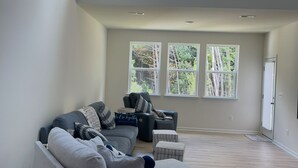 Ein geräumiges Wohnzimmer mit einem grauen Ecksofa, einem kleinen Hocker und einem dunklen Sessel. Drei Fenster bieten Blick auf Bäume.