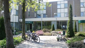 Un edificio con recepción, bicicletas estacionadas afuera y un jardín cuidado con arbustos y un banco.