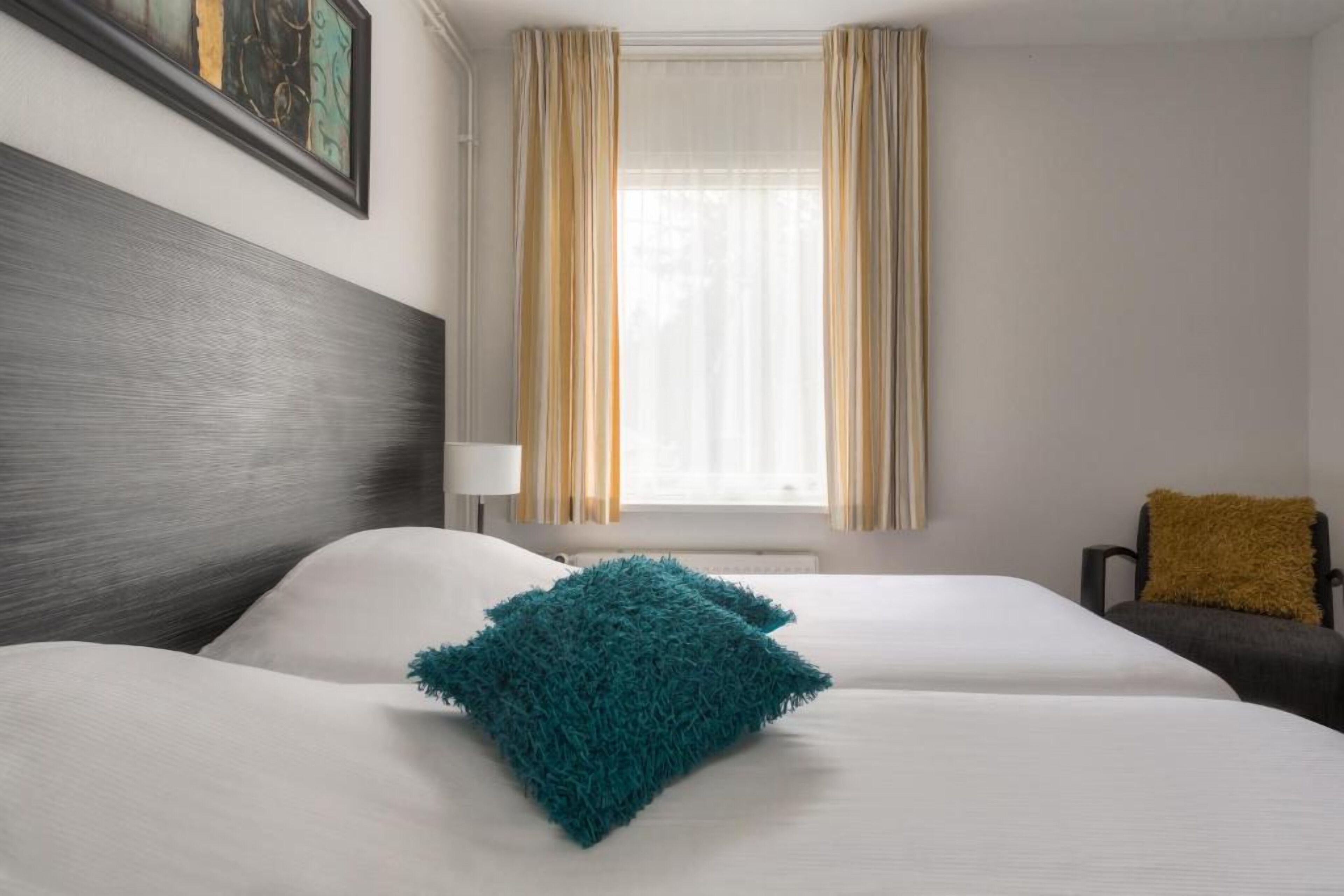 Une chambre d’hôtel avec un grand lit, une tête de lit, une fenêtre avec des rideaux, une chaise et un tableau au mur.