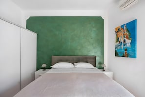 A bedroom with a green accent wall, a bed with white bedding, two bedside lamps, and a painting of a coastal scene. - Francy's Apartment with Balcony, Wi-Fi and Air Conditioning (Salerno)