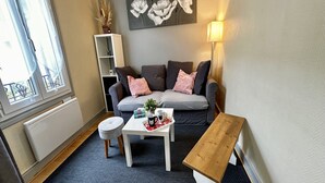 Un salón acogedor con un sofá gris, una mesa de centro blanca y un banco de madera. Se ve una ventana con vistas a un balcón.
