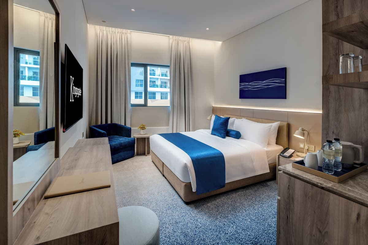 Ein Hotelzimmer mit einem Bett, einem Schreibtisch, einem Stuhl, einem Fernseher, einem Fenster mit Vorhängen, einem Gemälde und einem Nachttisch mit Lampe und Telefon.