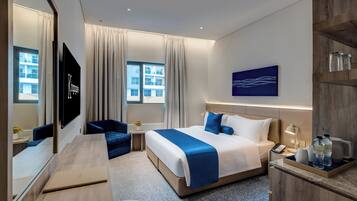 Habitación de hotel con cama, escritorio, silla, televisión, ventana con cortinas, un cuadro y mesita de noche con lámpara y teléfono.