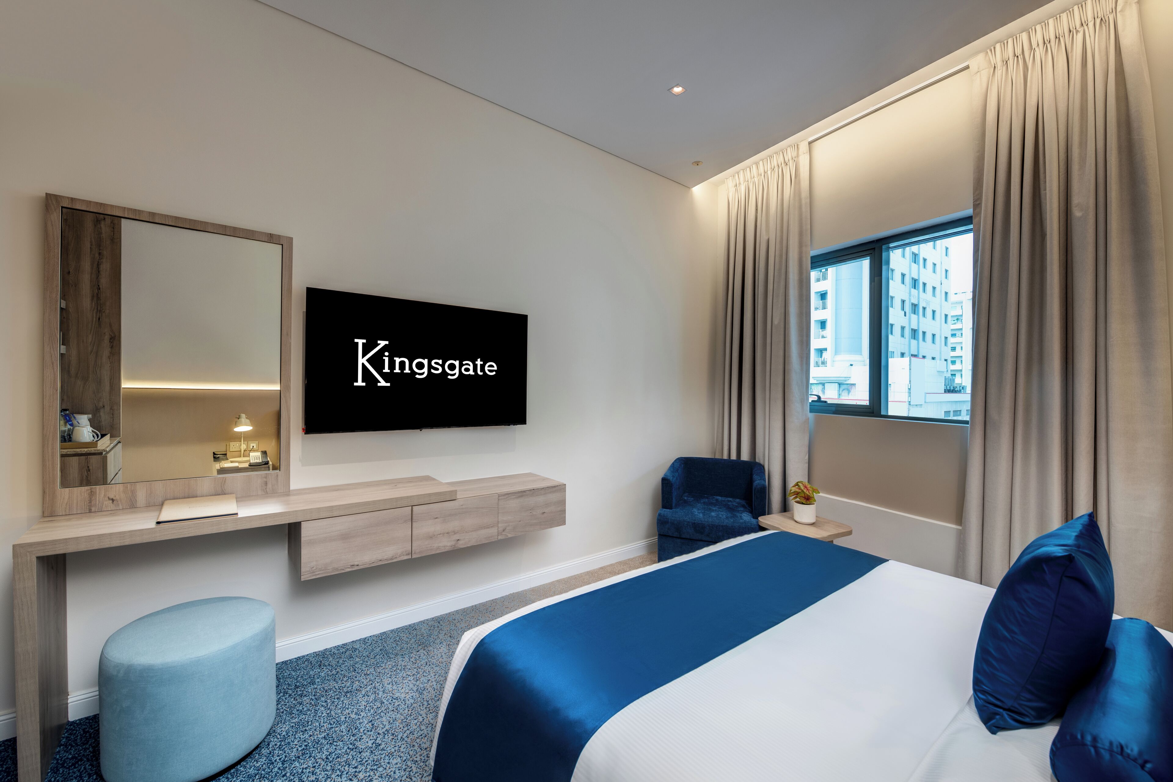 A hotel room with a bed, a TV mounted on the wall, a desk with a mirror, a chair, and a window with curtains.