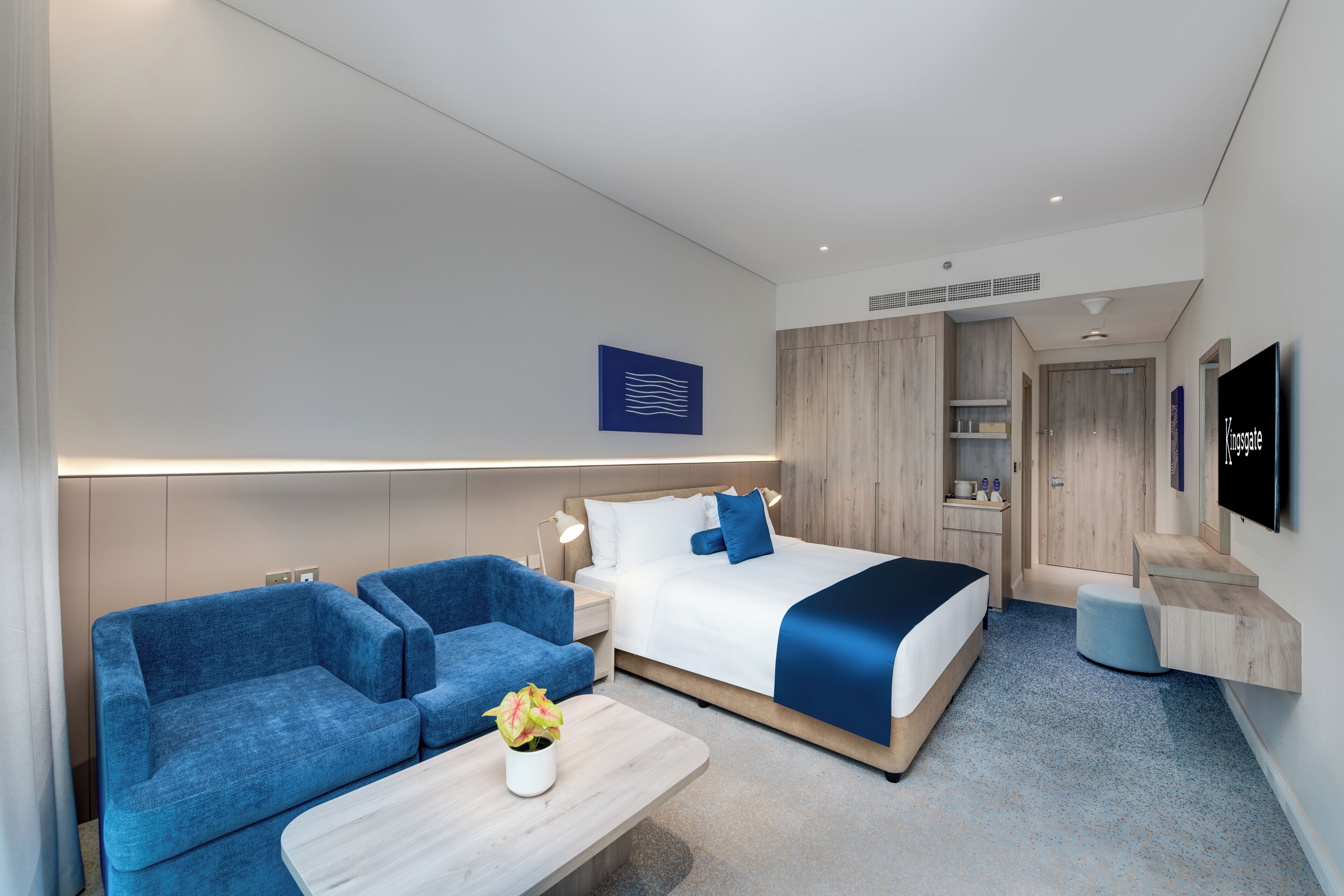 A modern hotel room with a bed, two blue sofas, a small table with a plant, a TV, and a wooden wardrobe.