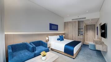 Una habitación de hotel moderna con una cama, dos sofás azules, una mesita con una planta, un televisor y un armario de madera.