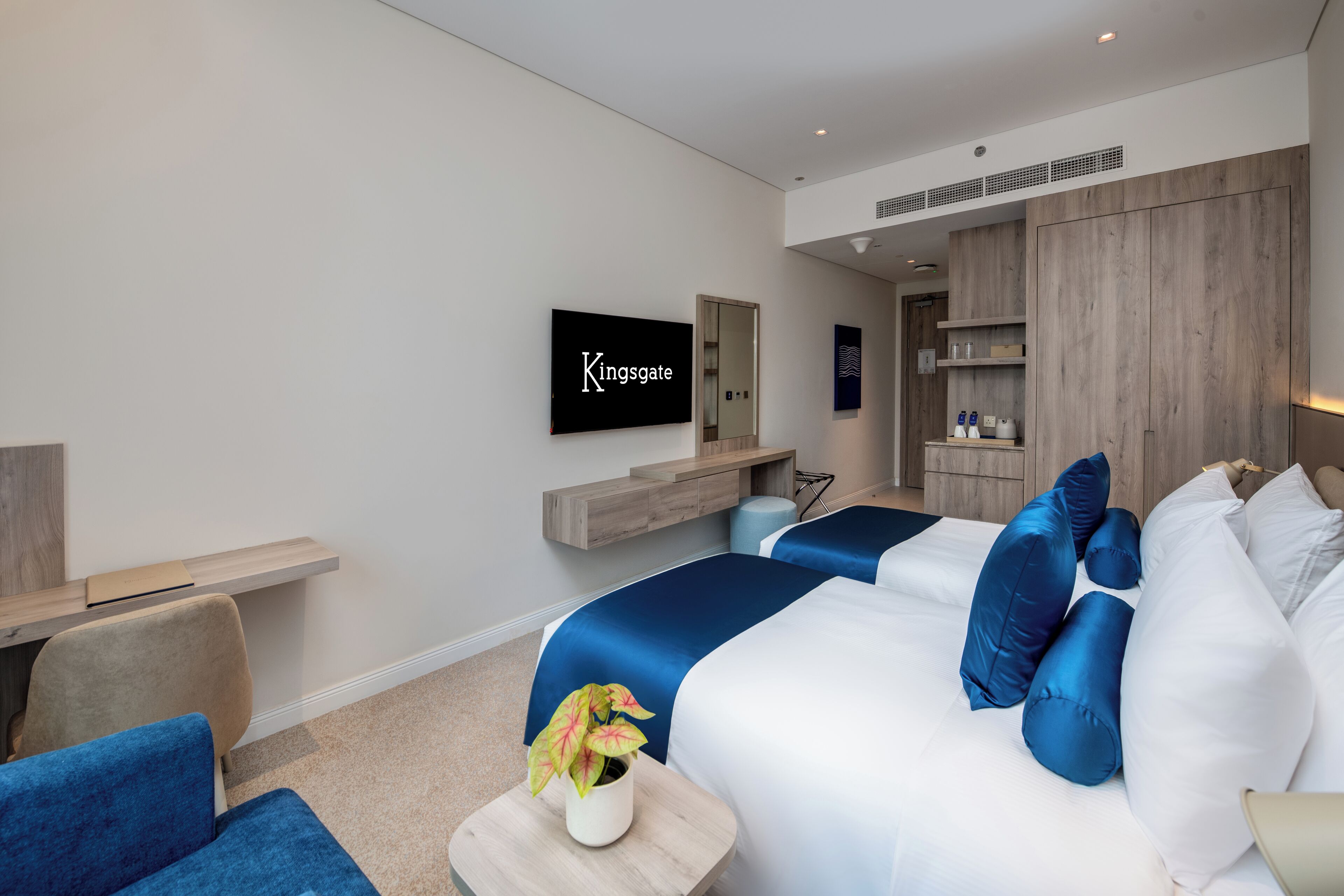 A modern hotel room with a flat-screen TV, a bed with blue and white bedding, a small table with a plant, and a wooden cabinet.