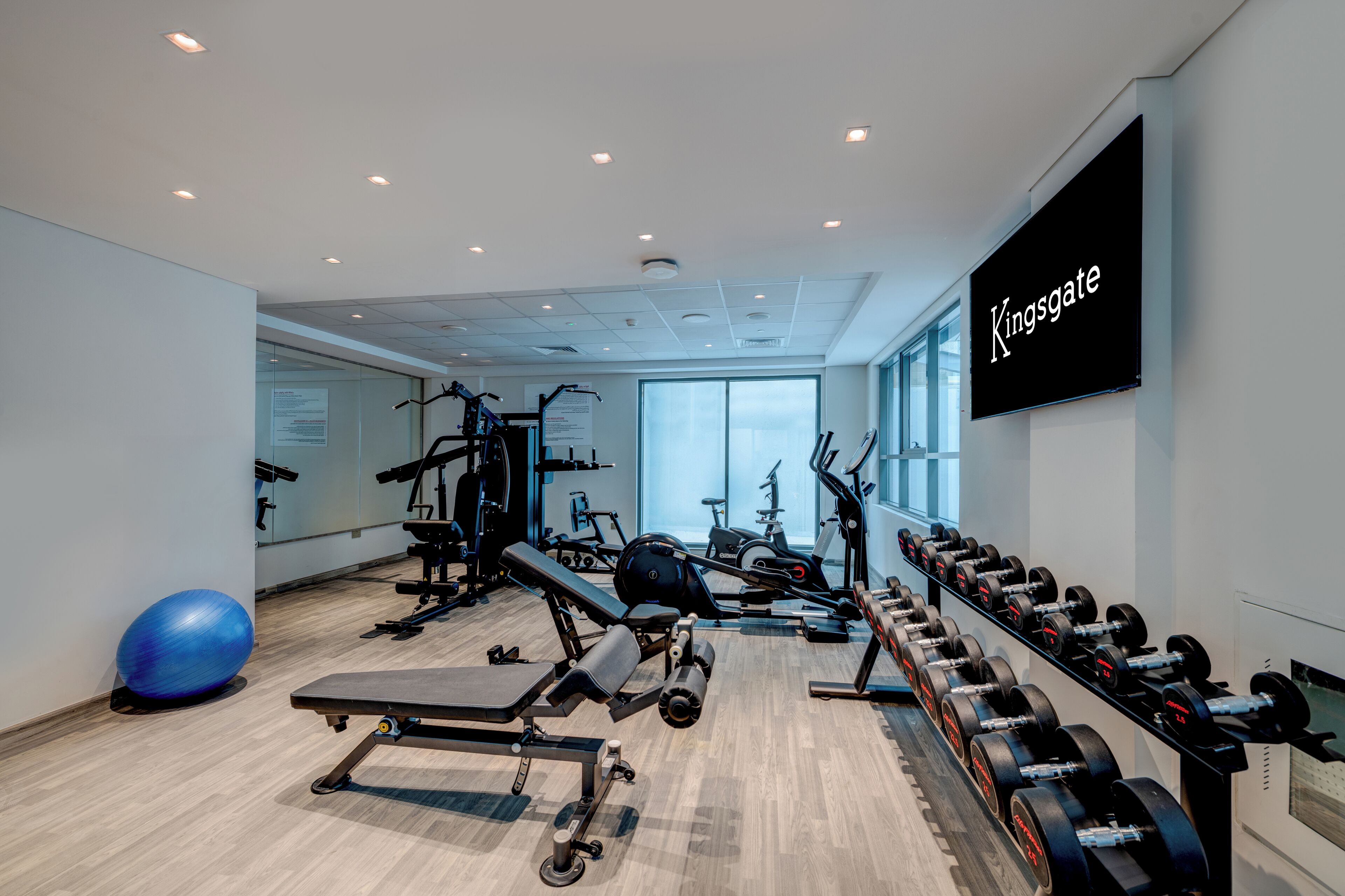 A modern gym with exercise equipment, a large screen displaying 'Kingsgate,' and large windows allowing natural light.
