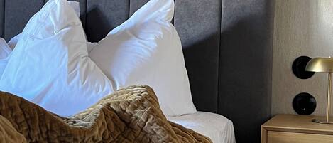 A neatly made bed with white pillows and a brown blanket, a grey upholstered headboard, and a bedside table with a lamp.