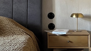 A neatly made bed with a dark headboard, a wooden nightstand with a lamp, and a wall with a textured finish. - hellofrom Tobacco Warehouse (Kavala)