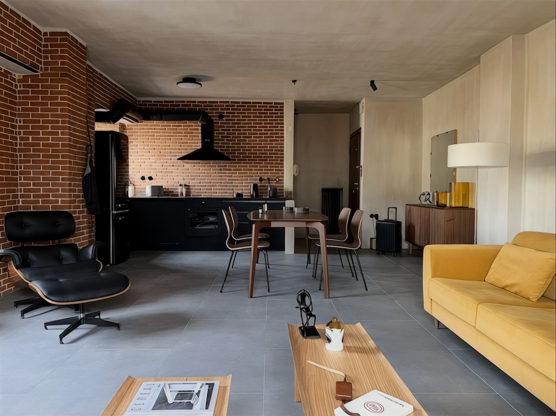 Ein modernes Wohnzimmer mit einem gelben Sofa, einem Essbereich mit Tisch und Stühlen, einer Küche mit Backsteinwand und schwarzen Geräten sowie einem schwarzen Sessel.