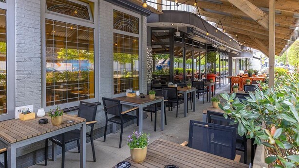 An outdoor cafe with wooden tables and chairs, large windows, and potted plants.