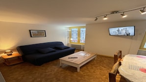 A living room with a dark blue sofa, a wooden coffee table, a small side table with a lamp, a TV mounted on the wall, and a window with a view of buildings. - Bright Apartment in Mont-Dore (Mont-Dore)