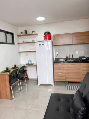 A compact kitchen with a refrigerator, wooden cabinets, a dining area with chairs, and a black sofa. - Moana Residencial - VSH (Ipojuca)