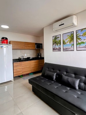 A modern kitchen with wooden cabinets, a white refrigerator, and a black countertop. There is a black leather sofa with cushions and a coffee machine on the counter. - Moana Residencial - VSH (Ipojuca)