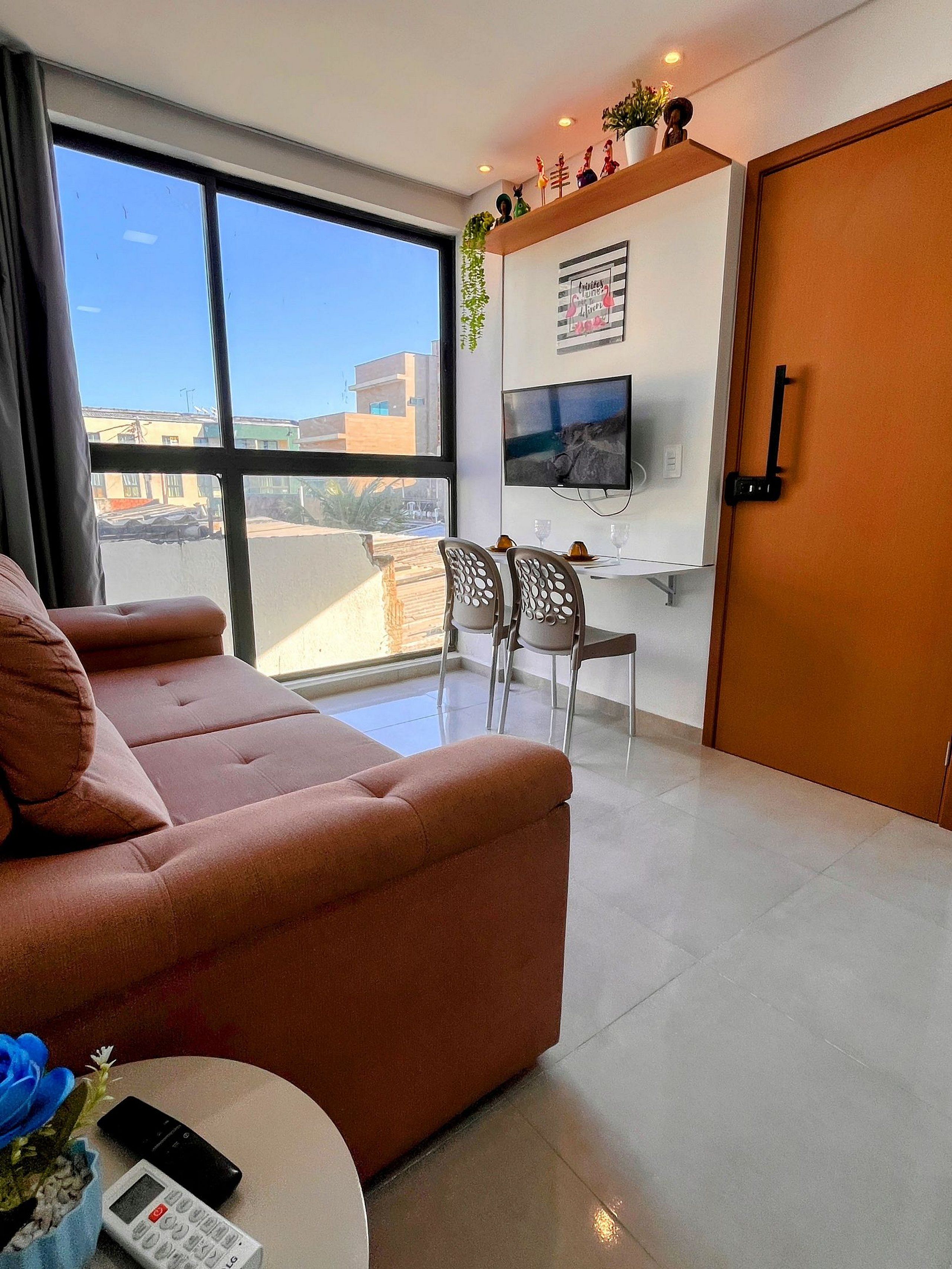 A modern living room with a sofa, a dining table with chairs, a TV mounted on the wall, and a large window offering a view of the city.