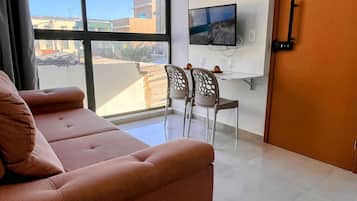 A modern living room with a sofa, a dining table with chairs, a TV mounted on the wall, and a large window offering a view of the city.