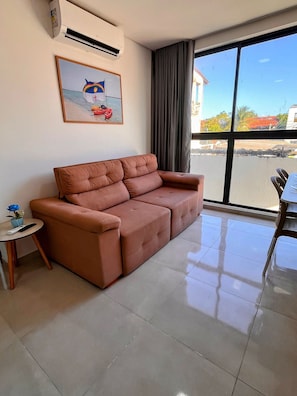 A living room with a sofa, a small table with a vase, and a painting on the wall. - Moana Residencial - VSH (Ipojuca)