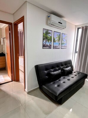 A modern living room with a black sofa, a wall-mounted air conditioner, and three framed pictures of tropical scenes. - Moana Residencial - VSH (Ipojuca)