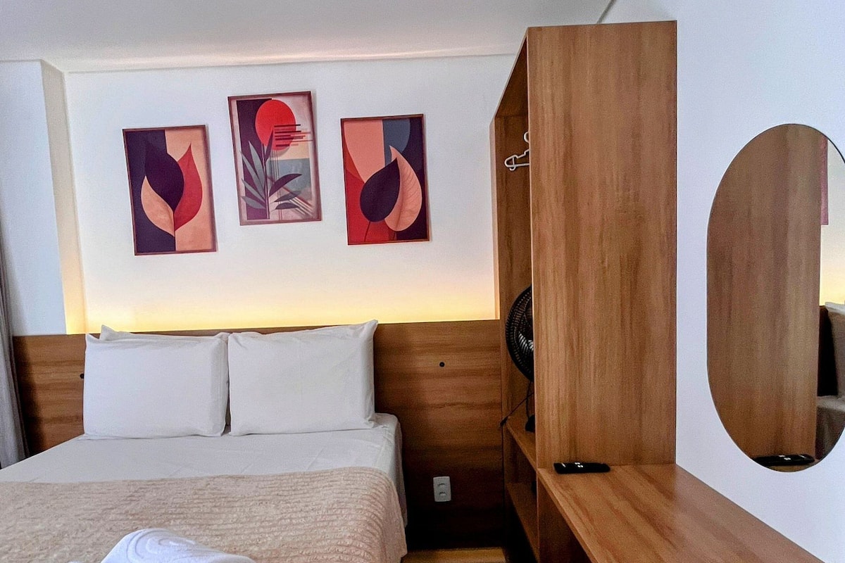A hotel room with a bed, a wooden nightstand, a mirror, and three abstract paintings on the wall.