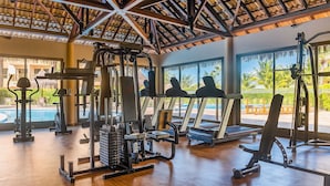 Un gimnasio con equipo de ejercicio, ventanas amplias y vistas a una piscina y palmeras.