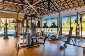 Uma academia com equipamentos de ginástica, janelas amplas e vista para uma piscina e palmeiras.