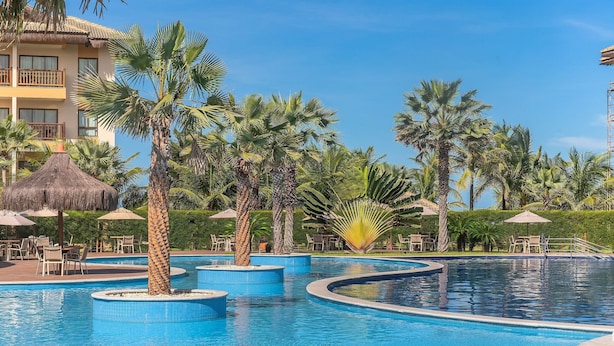 Un área de piscina en un complejo turístico con varias piscinas, palmeras y mobiliario exterior.