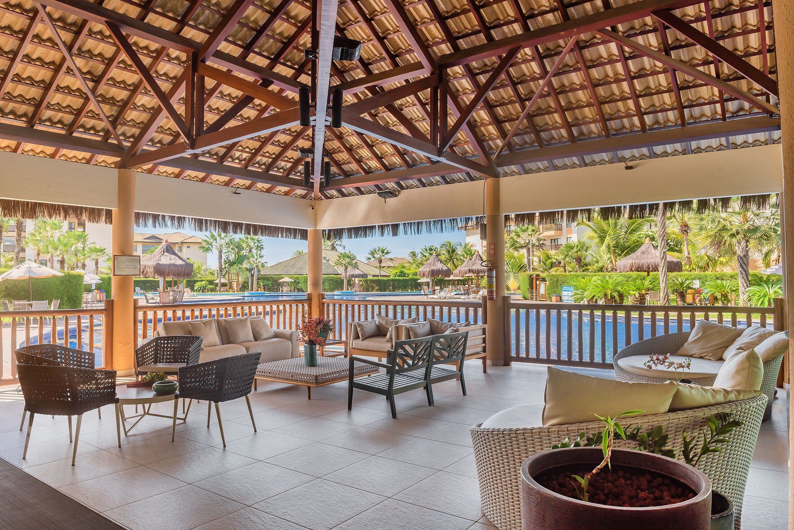 A covered outdoor lounge area with wicker furniture, a pool view, and a tropical garden backdrop.