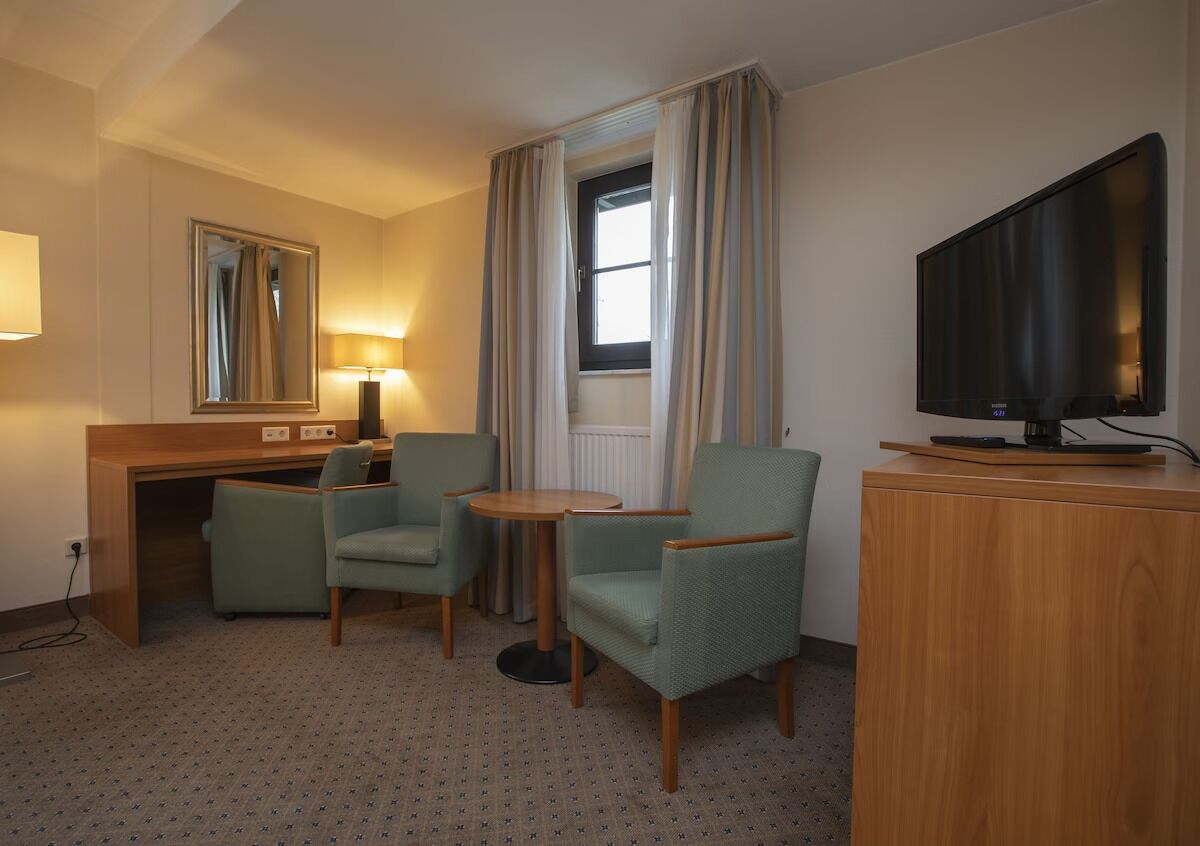 A hotel room with a wooden desk, two armchairs, a small round table, a television on a wooden cabinet, and a window with curtains.